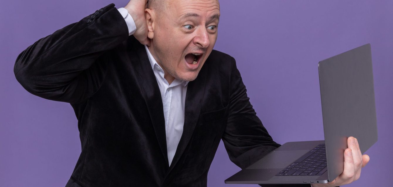 middle-aged-bald-man-suit-holding-laptop-looking-screen-screaming-being-confused-excited-standing-purple-wall-scaled-2560x1280